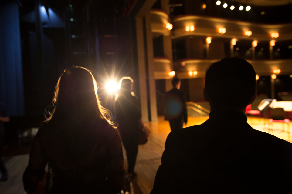 A silhouette of two students walking onto a well lit stage in a theatre.
